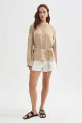 Blusa de poliéster color beige, con cuello alto, frunces en el escote y mangas largas con puños. Cuenta con un delicado lazo ajustable en la cintura y cierre frontal con botones.