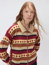 Campera de punto estampada de corte holgado, con cierre metálico frontal y cuello de corderito sintético en color tostado. Presenta un diseño de franjas horizontales con patrones geométricos en tonos bordó, mostaza, marrón y beige.