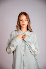 Camisa beige de corte amplio, con cuello clásico, cierre frontal con botones, mangas largas con puños abotonados y bordado floral en las mangas.