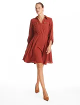 Vestido de crepe color marrón, con cuello solapa, escote en V y cintura elastizada. Presenta bolsillos simulados con cierres metálicos en el frente y mangas largas regulables con presilla y botón.