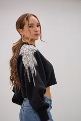 Sweater negro de corte cropped con detalle de apliques bordados en pedrería y lentejuelas plateadas sobre el hombro. Presenta mangas largas con puños ajustados y cuello redondo.