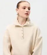Buzo de algodón color beige, con cuello polo y cierre frontal parcial con botones dorados. Presenta un diseño de corte holgado con mangas largas y puños acanalados.