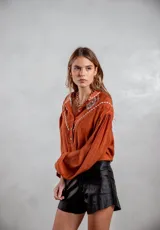 Camisa de tela texturizada color terracota, con mangas largas abullonadas y detalle de bordado blanco en el canesú delantero. Presenta cierre frontal con botones.