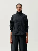 Chaqueta cortaviento negra, con cuello alto y cierre frontal. Presenta un diseño holgado y tejido técnico con textura arrugada.
