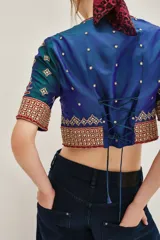Camisa de seda con diseño tipo chaqueta corta, de color azul petróleo con bordados decorativos en tonos dorados y rojos. Presenta mangas largas abullonadas de color crema y cierre frontal.