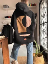 Campera tipo hoodie de textura tipo corderito en color negro, con diseño de collage abstracto en la espalda compuesto por formas geométricas en tonos marrón y rosado. Presenta capucha, mangas largas y un corte holgado.