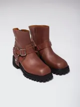 Bota corta de cuero color marrón, con punta cuadrada, suela track negra y estribo desmontable con hebilla metálica.