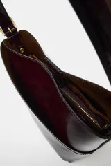 Bolso tipo saca de diseño asimétrico con acabado brillante, asa ajustable con hebilla lateral y cierre mediante botón imán.