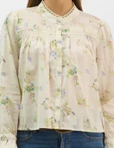 Blusa de algodón con estampado floral de flores silvestres sobre fondo claro. Presenta cuello redondo con detalle de puntilla, abotonadura frontal, canesú con frunces y mangas largas con puños elásticos decorados con puntilla.