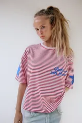 Camiseta oversize de rayas horizontales rojas y blancas, con cuello redondo y mangas cortas. Presenta un estampado de texto azul en el pecho y estrellas azules en las mangas.