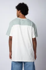 Remera de corte holgado con diseño color block, combinando paneles en color verde menta y blanco. Presenta un estampado frontal con la palabra 'Lucky' dentro de un óvalo y dibujos de flores con lentes de sol.