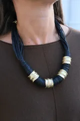 Collar de diseño artesanal compuesto por múltiples hilos de cordón negro entrelazados, intercalados con piezas cilíndricas de metal dorado con textura irregular y acabado rústico.