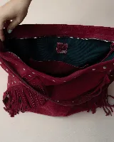 Cartera estilo hobo de gamuza color bordó, con flecos laterales y asa ancha decorada con tachas metálicas plateadas.