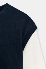 Prenda de manga larga blanca con un chaleco de tejido de punto azul marino superpuesto, creando un efecto de dos piezas. El chaleco es de cuello redondo y corte recto.