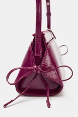 Bolso de hombro de material efecto acharolado en color fucsia, con diseño alargado, doble compartimento exterior y detalle de lazos en los laterales. Cuenta con doble asa de hombro ajustable y cierre superior con cremallera.