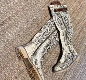 Botas altas de caña media con estampado de piel de serpiente en tonos beige, negro y blanco. Presentan una suela track de goma color caramelo, cierre lateral metálico y detalle de correa de cuero marrón en la parte superior de la caña.