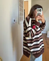 Sweater de punto oversized con diseño de rayas horizontales en tonos marrón, blanco y negro, cuello alto y mangas largas con hombros caídos. Cuenta con aberturas laterales en el dobladillo y una etiqueta roja decorativa en la manga.