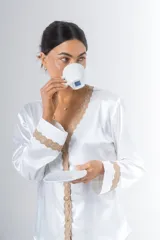 Pijama blanco de satén elastizado, compuesto por una camisa de manga larga con escote en V, abotonada adelante y con puntilla en escote y puños, y un pantalón recto con bolsillos laterales, elástico en la cintura y puntilla en el ruedo.