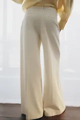 Pantalón wide leg de tiro medio confeccionado en hilatura de algodón, color beige, con trabillas en la cintura, bolsillos laterales ocultos y detalle de pliegues en el delantero. Cierre frontal con cremallera, botón y ganchos metálicos.