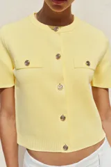 Chaqueta de punto color amarillo pálido, con cuello redondo, manga corta, cierre frontal con botones dorados y bolsillos de parche en el pecho.