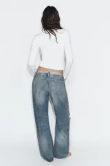 Jeans de tiro medio con diseño de cintura doblada, cinco bolsillos y cierre frontal con cremallera y botón metálico. Presenta un acabado desgastado con roturas en las rodillas y corte recto.