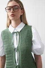 Chaleco tejido color verde musgo, con trama texturada, cuello a la base y cierre frontal con lazos. Presenta un ruedo con volado.