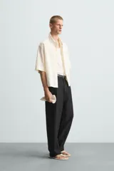 Camisa de corte relajado (relaxed fit) color crema o blanco roto, confeccionada en tejido con mezcla de algodón. Presenta un bordado sutil en relieve con patrón de ondas o líneas diagonales entrecruzadas. Tiene cuello solapa, manga corta y un bolsillo de plastrón en el pecho.