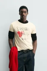 Camiseta de corte regular, color crema, con cuello redondo y ribetes en las mangas y cuello en color negro. Presenta un estampado frontal con un corazón rojo y texto en negro que dice "Everything beats for something".