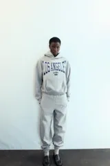 Sudadera gris con capucha y manga larga. Presenta un bolsillo canguro en la parte delantera y un estampado en azul marino con el texto "LOS ANGELES CALIFORNIA EST. 1975". Los acabados son de rib.