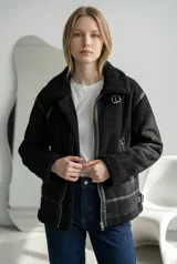 Campera tipo gamulán negra con interior y detalles de cuello y puños en corderito sintético. Presenta cierre frontal metálico, correas ajustables con hebillas en el cuello y en los laterales, y ribetes de cuero sintético.