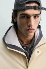 Parka de tejido técnico color beige claro, corte relajado (relaxed fit), con cuello subido, cierre frontal oculto por solapa con botones a presión y bolsillos de solapa en cadera.