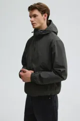 Campera rompevientos de nylon color verde oscuro, con capucha ajustable con cordón, cierre frontal y puños elásticos. Presenta bolsillos laterales y un bolsillo interior, todos con cierre.