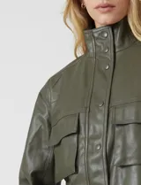 Chaqueta corta de eco cuero color tostado, con cuello alto, cierre frontal con botones a presión y bolsillos de solapa en el pecho. Tiene forro interno a tono.