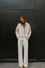 Trench corta de color beige, con cuello solapa, mangas largas con trabillas en los hombros, bolsillos delanteros de vivo y cierre frontal cruzado con botones a presión.