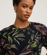 Blusa de manga corta y escote redondo, confeccionada en viscolycra con un estampado de hojas tropicales en tonos verdes, rojizos y crema sobre fondo negro.