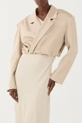 Blazer corto y cruzado, de color beige, con hombros estructurados, solapas y cierre lateral. El largo termina en la cintura, con un frunce que le da forma, confeccionado en un tejido de leve brillo.