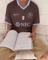 Camiseta de fútbol color marrón con cuello en V y detalles en celeste, inspirada en el Napoli F.C., con diseño de mangas cortas y sponsors impresos en el frente.