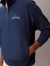 Buzo Calvin Klein de color azul marino, confeccionado en mezcla de algodón y poliéster. Cuenta con cuello alto, cierre de media cremallera y manga larga con hombros caídos. Presenta puños y dobladillo acanalados, y el logo de la marca bordado en punto de cadeneta en el pecho.