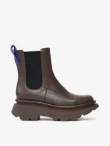 Bota tipo Chelsea de cuero de oveja color marrón, con paneles elásticos laterales negros y suela chunky dentada. Incluye un tirador trasero con detalle tejido en azul.