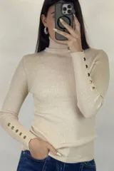 Polera de hilo blanca de cuello alto y manga larga, con detalle de botones dorados en los puños. Tejido de punto acanalado ajustado al cuerpo.
