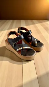 Sandalias de cuero color marrón oscuro con plataforma y taco de 5 cm. Tienen tiras cruzadas en el empeine con apliques metálicos decorativos y un dije colgante en forma de triángulo, además de una tira con cierre ajustable en el talón.