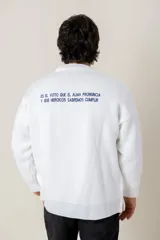 Buzo de tejido de punto color blanco, de corte holgado y cuello redondo. Presenta un bordado central en hilo dorado con el escudo nacional y un pequeño detalle bordado en la manga derecha con los colores de la bandera uruguaya.