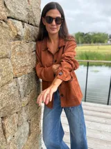 Blazer de gamuza sintética color terracota, con cierre frontal de botones y detalles de bordado a mano en contraste en los bordes, cuello y bolsillos. Presenta un diseño suave y flexible con mangas largas.