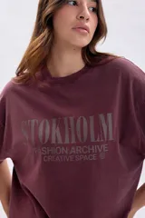 Remera de manga corta con corte boxy, color bordó con efecto de teñido especial. Presenta una estampa frontal con el texto 'STOKHOLM FASHION ARCHIVE CREATIVE SPACE' en tono oscuro.
