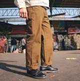 Pantalón de trabajo estilo cargo color marrón, confeccionado en resistente tela de algodón. Presenta un diseño de cinco bolsillos, incluyendo compartimentos utilitarios, cierre frontal con botón y trabillas reforzadas para cinturón.