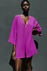 Mono corto de color fucsia, con escote en pico delantero y en la espalda, mangas anchas y largas, y detalle de fornitura dorada en el escote delantero con tiras finas para atar en la espalda.
