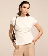 Blusa de manga corta color blanco hueso, con cuello redondo y un detalle de cruce o drapeado asimétrico en la parte delantera a la altura de la cintura. La tela presenta una textura sutil.