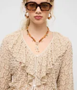 Blusa tipo bata de encaje texturizado color beige, con escote en V, mangas largas y volados aplicados en el escote. Presenta cierre frontal con botones y lazo de ajuste en la cintura.