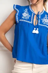 Blusa blanca de viscosa con bordados negros en contraste. Presenta escote en V con lazo y borlas, mangas cortas tipo volado y detalles bordados de flores y motivos geométricos en el pecho y hombros.