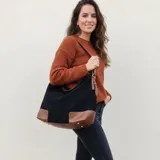 Bolso tipo hobo grande, de color negro con base y detalles en eco cuero marrón. Cuenta con una correa larga para el hombro y herrajes metálicos dorados.